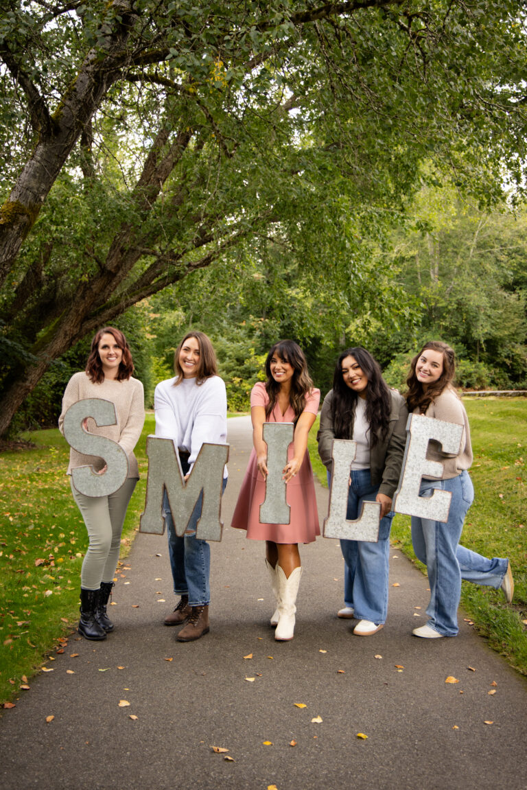 Five women holding large letters that spell SMILE on a tree-lined path