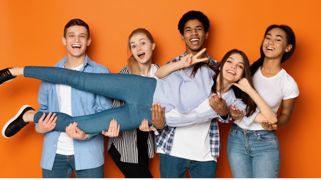 Group of five smiling teens lifting one friend horizontally on orange backdrop