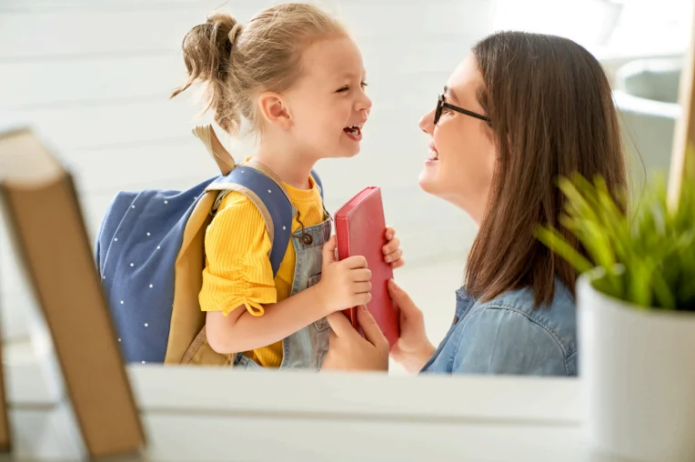 Smiling mother holding a red book talking with her young daughter wearing a backpack